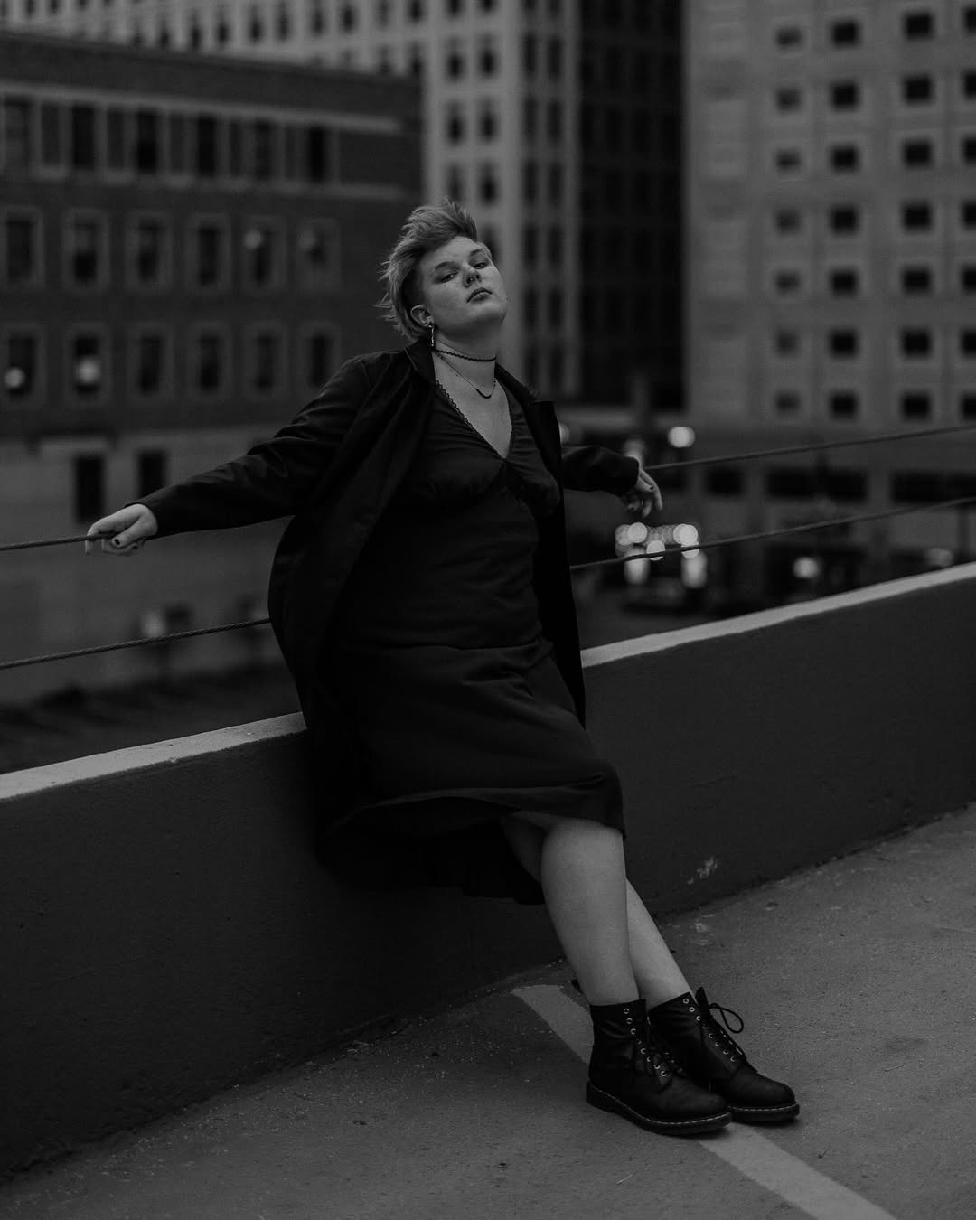 b/w photo of amara in a dress and docs leaning backwards over ledge on the top of a building