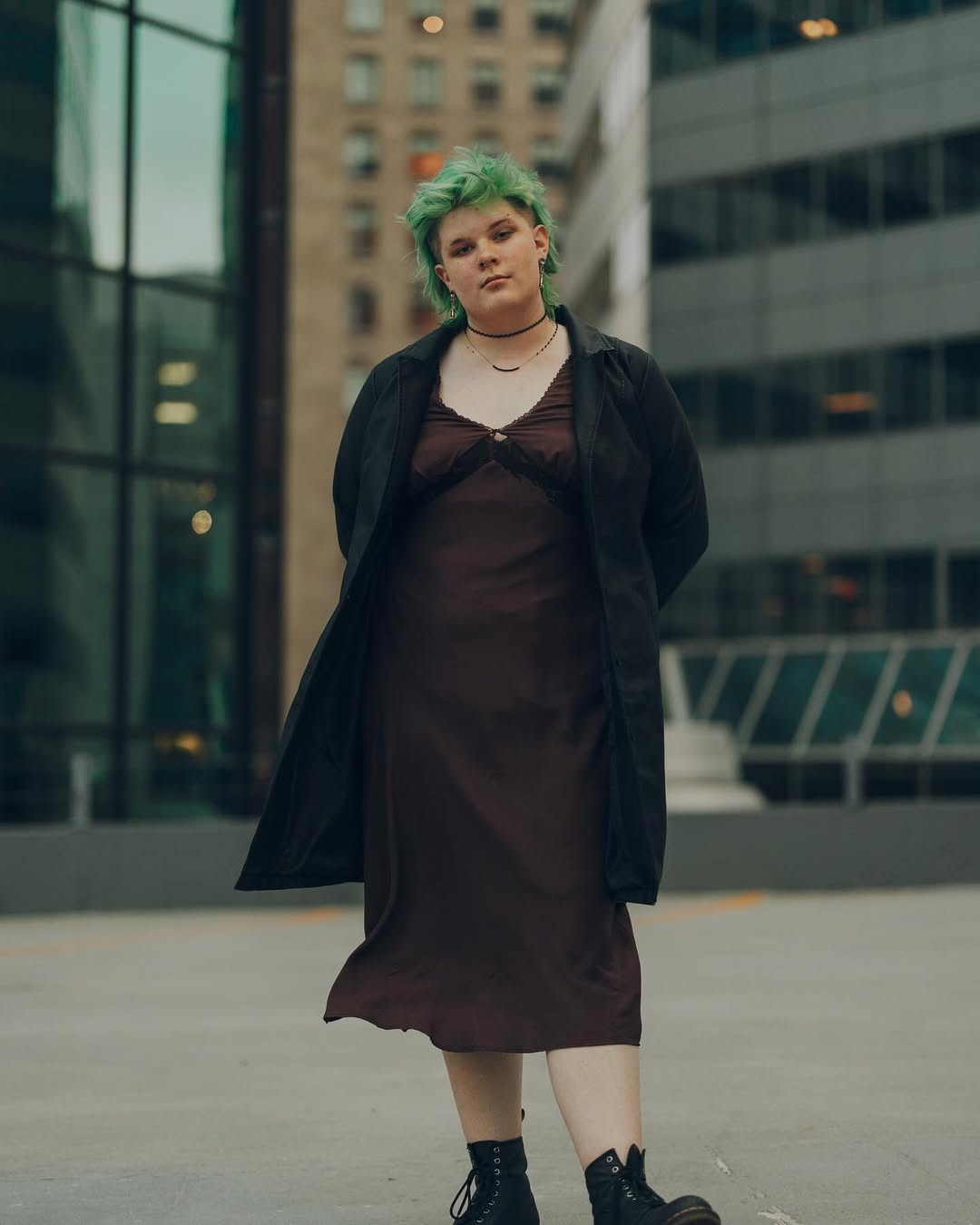 photo of amara with dress and leather trench coat standing on a building with one foot kicked in front